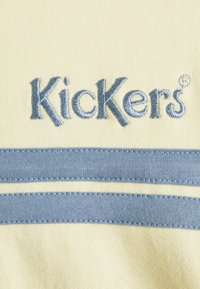 Kickers-logo geborduurd in blauwe draad op een lichtgele stof met daaronder twee horizontale blauwe stoffen strepen.