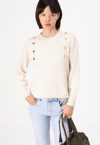 Cream knit sweater with round neckline, featuring two rows of metallic buttons on each shoulder. Worn with light wash jeans and a textured bag.