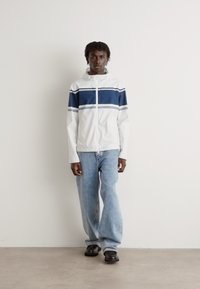 Hvid jakke med blå vandrette striber, lynlås foran, parret med lyseblå jeans og sorte sko, sat mod en neutral baggrund.