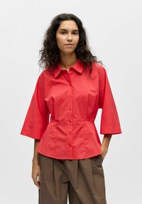 Femme aux longs cheveux bouclés portant une chemise rouge à boutons à manches larges et un pantalon plissé marron, debout devant un fond uni.