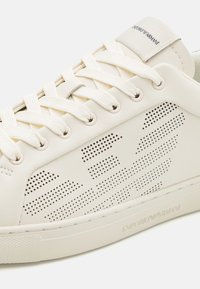 Vita lädersneakers med perforerade detaljer och platta bomullssnören. Diskret branding på tungan och sidan, med en texturerad yttersula.