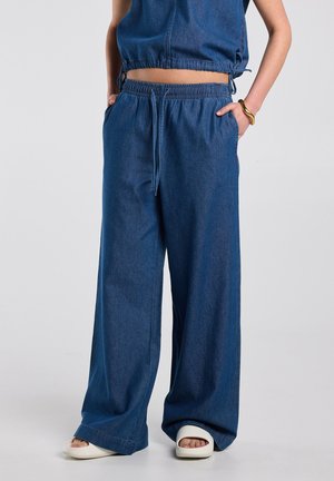 Persoon die blauwe wijde spijkerbroek en cropped mouwloze top draagt, met handen in de zakken, witte slip-on sandalen en een gouden armband.