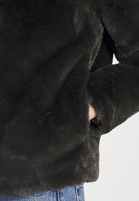 Chaqueta de piel sintética negra con una textura suave, que presenta un corte holgado y bolsillos metidos, combinada con una capa de ropa oscura debajo.