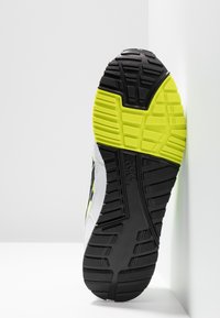 Suola della scarpa in gomma nera, con motivi di trazione profondi e accenti gialli brillanti. La parte superiore è realizzata in materiale sintetico bianco e nero.