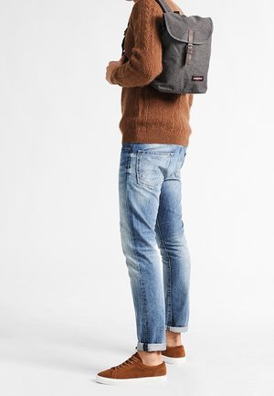 Harmaa kangasreppu läpällä ja solkikiinnityksellä, puettuna ruskean neuleen päälle, yhdistetty vaaleansinisiin farkkuihin ja ruskeisiin tennareihin.