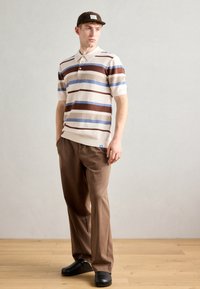Lys beige polo med brune og blå striper, korte ermer og krage. Brune bukser med avslappet passform og svarte klip.
