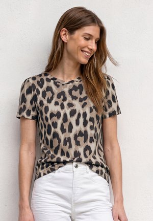 Kvinde med langt brunt hår iført en kortærmet leopardprint skjorte og hvide bukser, smilende og kigger ned til siden.