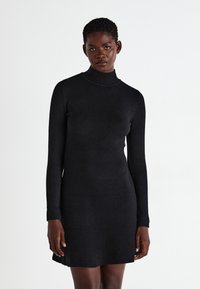 Fekete garbós ruha hosszú ujjakkal, testre simuló szabás, sima textúra és térd alatti hosszúság. A modell egy egyszínű háttér előtt áll.