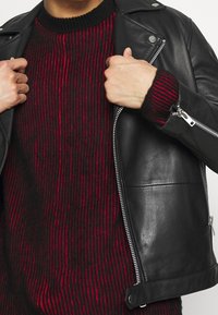 Veste de motard en cuir noir avec fermeture éclair argentée et détails à boutons-pression, portée sur un pull en tricot rayé rouge et noir avec une texture côtelée.