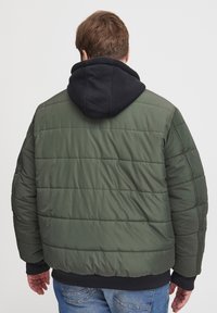 Grønn pufferjakke med horisontale sømmer, svart hette og ribbestrikkede mansjetter. Stoffet ser glatt og litt skinnende ut. Uformell design.