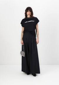 Melna T-krekls ar 'KARL LAGERFELD' logo, apvienots ar augsto vidukli melnām plisētām biksēm un mazu sudraba rokassomu. Spīdīgas melnas kurpes.