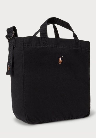Sac en toile noir fabriqué en tissu durable, avec des poignées doubles et une sangle amovible. Détail de logo brodé à l'avant.