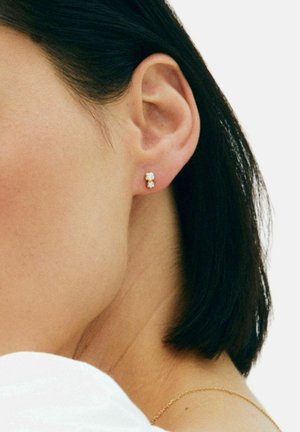 Žensko uho s kratkom tamnom kosom, nosi male naušnice s dvostrukim kamenom i tanku zlatnu ogrlicu na svijetloj pozadini.