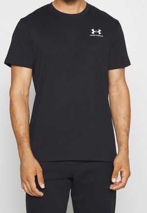 Mann trägt schlichtes schwarzes Under Armour T-Shirt mit kurzen Ärmeln und weißem Logo auf der linken Brust, kombiniert mit schwarzen Hosen, vor weißem Hintergrund.