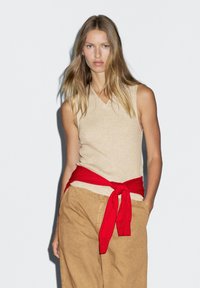 Top senza maniche beige in maglia con scollatura a V, abbinato a pantaloni larghi color sabbia. Una cintura rossa è annodata in vita, aggiungendo contrasto.