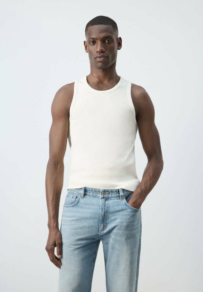 Homme portant un débardeur blanc sans manches et un jean bleu clair, debout avec une main dans la poche sur un fond clair uni.
