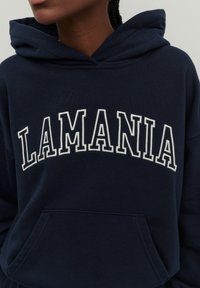Tamsiai mėlynas džemperis su kišene priekyje ir užtrauktuku ant galvos. „LAMANIA“ didžiausiomis baltomis raidėmis išlenktai parašyta ant krūtinės.