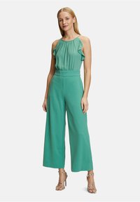 Grön jumpsuit med en volangkrage och vida ben, tillverkad av lättviktig tyg. Matcher med mönstrade, strap-on högklackade skor.