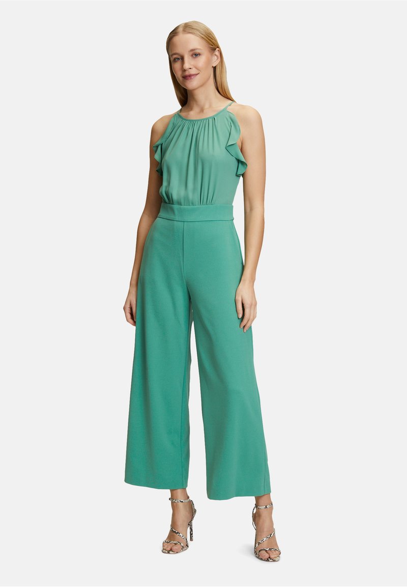 Grön jumpsuit med en volangkrage och vida ben, tillverkad av lättviktig tyg. Matcher med mönstrade, strap-on högklackade skor.