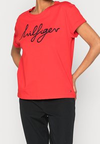 Red cotton t-shirt with a relaxed fit, featuring a black embroidered "hilfiger" logo. Short sleeves and a round neck design.