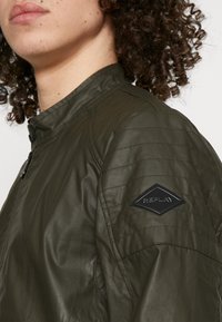 Olivgrön bomberjacka med hög krage, quiltade axelpaneler och en svart diamantlogotyp på vänster ärm. Slät textur.