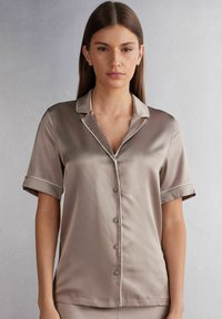 Silkeskjorta med kort ärm i taupe med klassisk krage, kontrasterande vit piping och knappstängning fram. Slät textur med avslappnad passform.