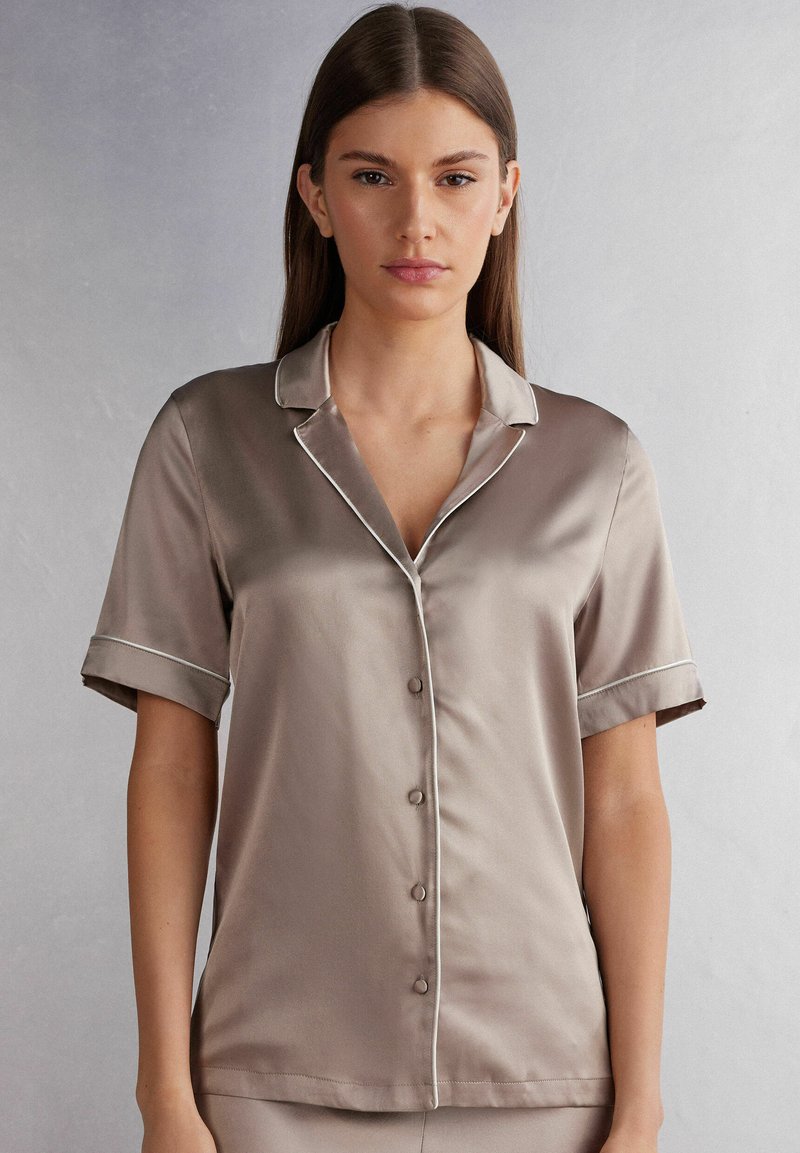 Silkeskjorta med kort ärm i taupe med klassisk krage, kontrasterande vit piping och knappstängning fram. Slät textur med avslappnad passform.
