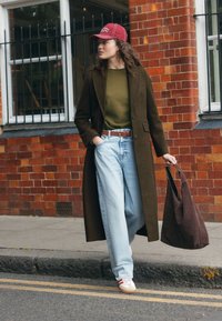 Manteau en laine marron sur un pull vert, jean large bleu clair, et baskets beige avec des rayures rouges. Sac cabas marron, cadre urbain décontracté.