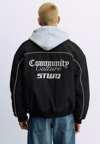 Zwarte jas met grijze capuchon, voorzien van geborduurde tekst: "Community Culture STWD." Inclusief witte biezen en geribbelde manchetten. Casual pasvorm.