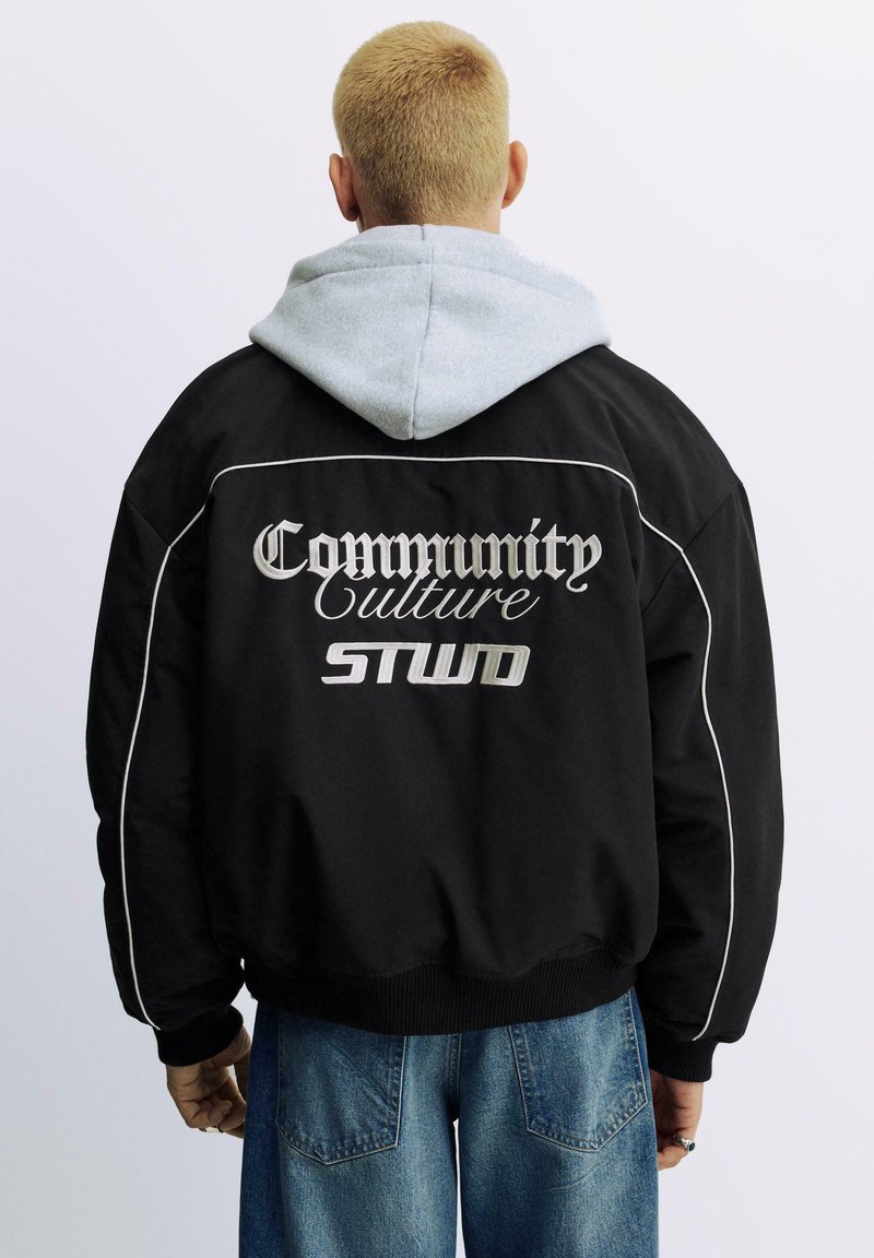 Zwarte jas met grijze capuchon, voorzien van geborduurde tekst: "Community Culture STWD." Inclusief witte biezen en geribbelde manchetten. Casual pasvorm.