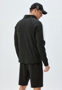 Chaqueta deportiva negra de mangas largas con detalles blancos, combinada con pantalones cortos negros, tejido mate con una textura suave.