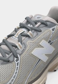 Κοντινή λήψη γκρι και μπεζ αθλητικού παπουτσιού New Balance που δείχνει υφή από δίχτυ, γκρι κορδόνια και το λογότυπο "N" στο πλάι.