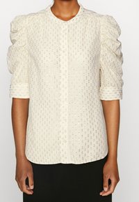 Blusa corta a maniche corte in tessuto crema con motivo a pois dorati. Presenta una chiusura con bottoni e spalle arricciate per una maggiore texture.