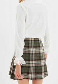 Valkoinen paita, jossa on pitsikoristeet hihoissa, yhdistetty vihreän, ruskean ja beige-ruudullisen hameen kanssa. Pehmeä kangas ja levenevä malli.