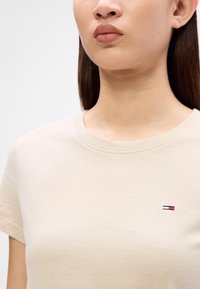 Beige bomull T-skjorte med korte ermer, rund hals og en liten brodert logo med røde, hvite og blå striper på brystet.