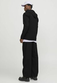 Schwarzer Kapuzenpullover mit langen Ärmeln, kombiniert mit weiten schwarzen Hosen und schwarzen Stiefeln; eine weiße Mütze bildet einen Kontrast zu dem dunklen Outfit.