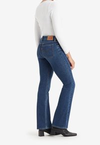 Femme portant un jean bleu droit, des bottes noires à talons et un haut blanc à manches longues, debout de profil sur un fond blanc.