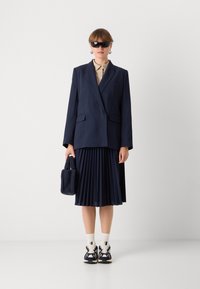 Námořnický pruhovan�ý oversize blazer, skládací sukně po kolena, béžová košile s límečkem, černé sluneční brýle, texturovaná námořnická kabelka a tenisky.