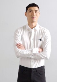 Chemise blanche à manches longues, avec un col pointu et un détail logo noir, associée à un pantalon noir. Tissu lisse, design ajusté.