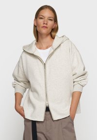 Chaqueta con capucha de cremallera color gris claro, de textura suave, con puños acanalados y un ajuste relajado. Llevada sobre una camisa blanca y pantalones beige.