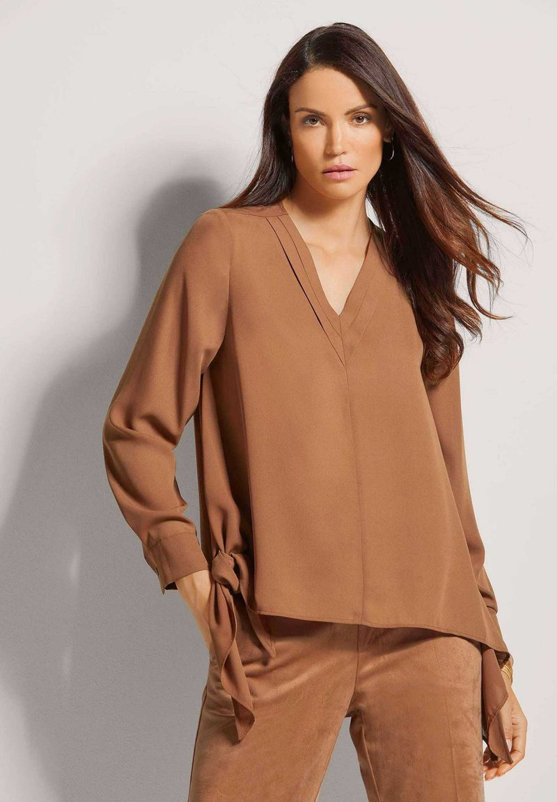 Braune Bluse mit langen Ärmeln, V-Ausschnitt und Bindebändern an der Taille. Glattes, leichtes Material mit dezenten Faltendetails.