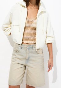 Hellbeige, taillierte Jacke mit Zugband, getragen über einem goldenen Strickoberteil, kombiniert mit hellblauen Jeansshorts, die einen lässigen Sommerlook präsentieren.