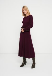 Bordeauxrood gebreide jurk met lange mouwen, een nauwsluitend lijfje en een uitlopende rok, gecombineerd met een zwarte clutch en donkerkleurige laarzen met hak tot op de knie.