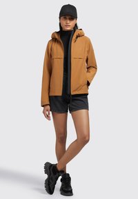 Brown, lightweight jacket with hood and front pockets, worn over a black turtleneck and dark shorts, paired with black sneakers.