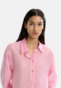 Różowa lniana koszula z guzikami i kołnierzykiem, o lekkiej fakturze, luźnym kroju i srebrnych guzikach.