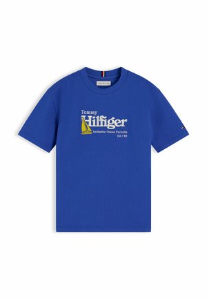 Drenge iført en blå Tommy Hilfiger T-shirt, beige vidde bukser og beige sneakers, stående med den ene hånd i lommen mod en hvid baggrund.