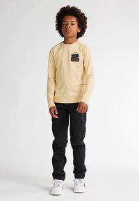 Pitkähihainen keltainen puuvillapaita, jossa on puun kuvio, yhdistettynä mustiin cargo-housuihin ja valkoisiin lenkkareihin. Asu on väljä ja rento.