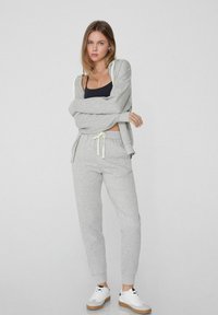 Lichtgrijze hoodie met koordjes, gecombineerd met bijpassende joggers. Model draagt een zwarte tanktop en witte sneakers. Glad, casual stof.