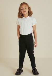 Witte polo met korte mouwen en drie knopen, gecombineerd met zwarte strakke broek en glanzende zwarte schoenen met een dikke zool.