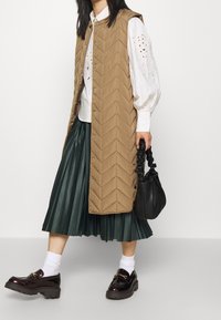 Hnědý prošívaný dlouhý vesta s chevron vzorem, bílá blůza se vzory, zelená skládací sukně, černá kulatá kabelka a lesklé černé mokasíny s mohutnými podrážkami.